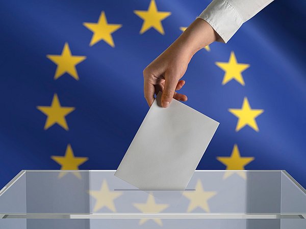 Eine Hand, die einen Stimmzettel in eine Wahlurne wirft. Im Hintergrund eine EU-Flagge.